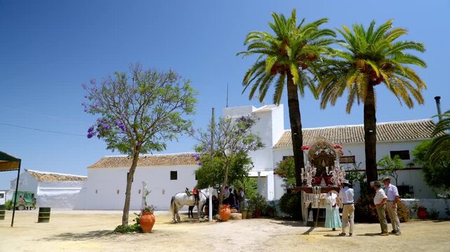 a Traditional Roc&iacute;o courtyard under palm trees with decorated shrine and people gathered in warm Andalusian sunlight during pilgrimage season