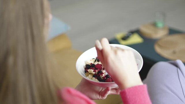 Woman mixes and prepares to eat a nutritious breakfast bowl with skyr and forest fruits on the sofa, emphasizing health-conscious lifestyle, morning wellness, and balanced diet at home.