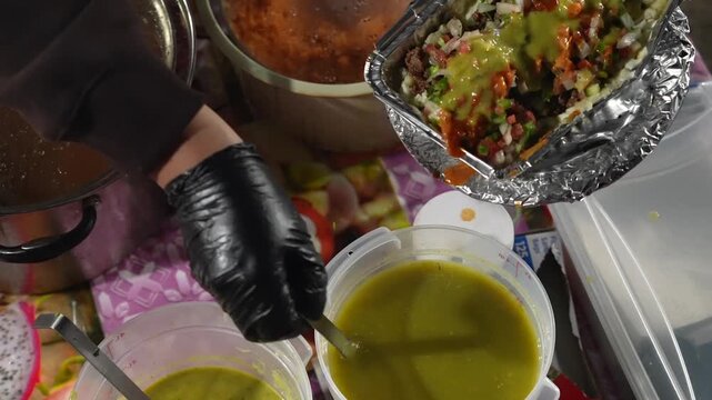 Close up footage of papa loca held by a vendor as green sauce is ladled and spread over the toppings. Slow camera movement highlights sauce texture, coverage, and street cuisine presentation.