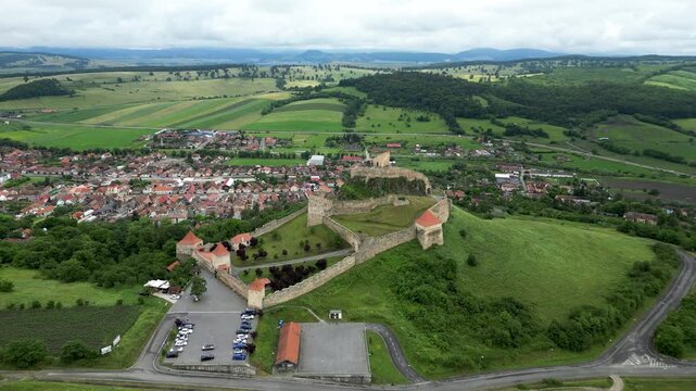 Aerial drone footage flying forward toward Rupea Fortress in Transylvania, with the town of Rupea visible behind the citadel. Medieval landmark, scenic countryside, travel and heritage concept.