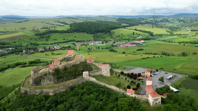 Aerial drone footage pulling backward from Rupea Fortress in Transylvania, revealing medieval stone walls, surrounding green hills and rural countryside. Cinematic historic landmark, travel and herita