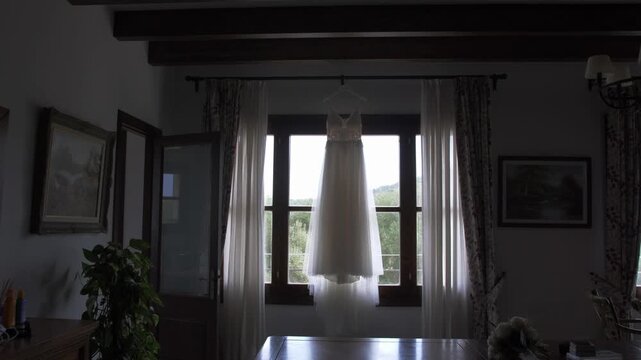Wedding dress hanging in front of a window inside a traditional Mallorca finca with soft daylight and slow camera movement