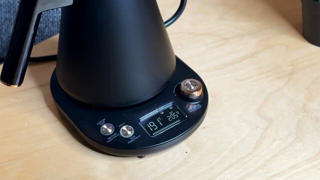 Close-up of a hand pressing the start button on a temperature-control electric kettle base, showing 191&deg;F and 205&deg;F set point on display, indoors on a wooden table.