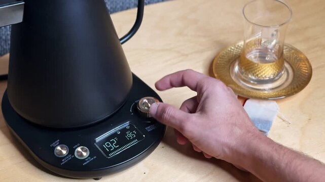 Close-up hand turning dial on a temperature-control electric kettle base, digital display at 192&deg;F, with tea bag and empty glass cup on saucer on a wooden table indoors.