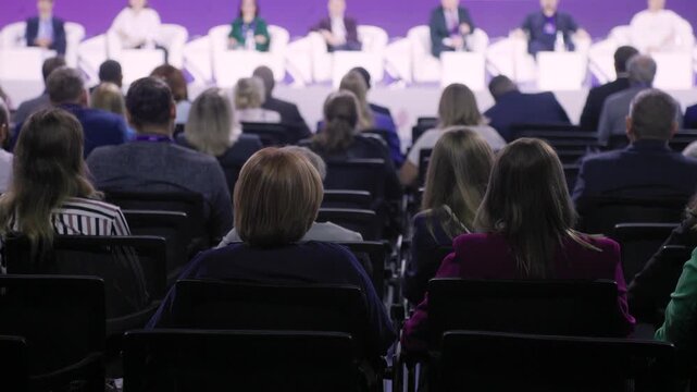 conference delegates seated near stage, front rows show professionals exchanging glances and notes during lively panel discussion, varied attire, collaborative