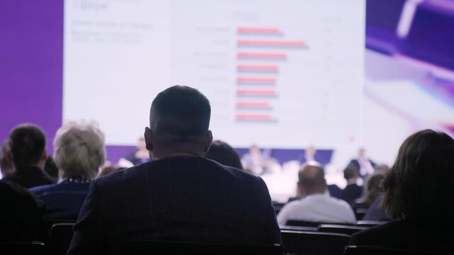 conference audience watching analytical presentation rows of seated professionals dim auditorium large projection screen with bar chart rear silhouette focused