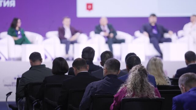 panelists on stage with audience attentive delegates seated rows purple backdrop white armchairs female speaker green professional exchange QA atmosphere