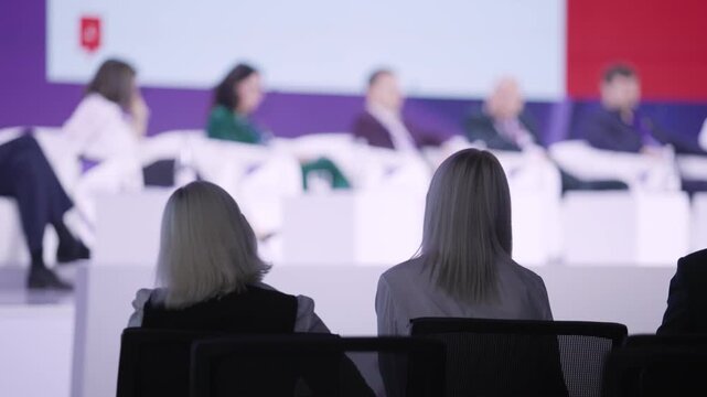 two delegates observing panel debate frontrow seats blurred speakers on stage focused notetaking formal conference signage reflective concentration