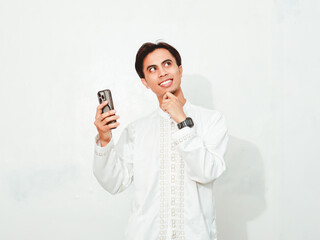 Fototapeta premium Asian Muslim man smiling while holding smartphone and looking up with thinking gesture, isolated on white background for technology and lifestyle advertising