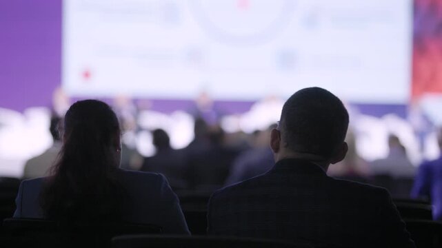 silhouetted couple watching illuminated screen backlit attendees dark auditorium blurred speakers data visuals projected reflective evening session corporate