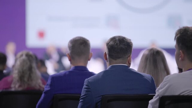 audience seated at conference listening attentively, business professionals facing stage with blurred presenter and projection screen, purple backdrop, rows