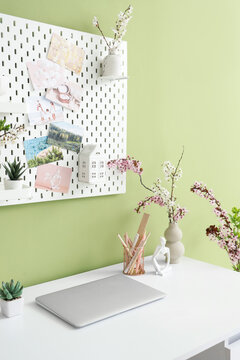 Workplace with laptop, blossoming branches and pegboard on green wall in office
