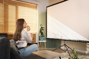 Young woman with popcorn and modern projector sitting on sofa in stylish living room