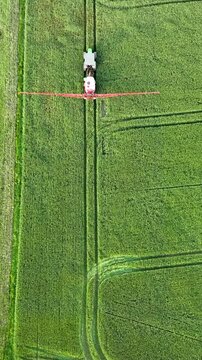 Vertical aerial shot of a tractor with a boom sprayer applying pesticides or fertilizers to a lush green crop field, ideal for agricultural and agribusiness content.