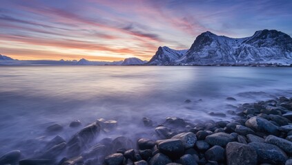 Calm winter dawn in Lofoten, Norway, with gentle waves on rounded stones and snow-capped mountains beneath a vibrant sky