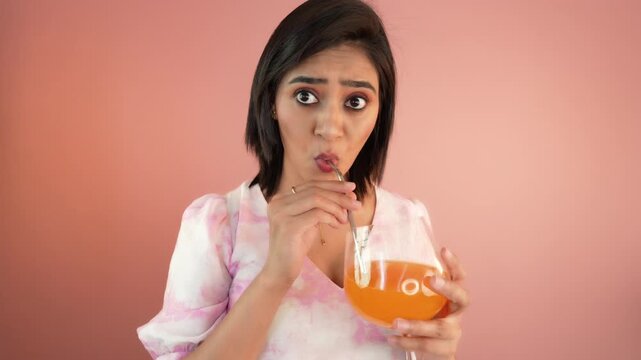 Close up portrait of surprised pretty young Indian woman lady holding glass sipping orange juice staring at camera with amazed face expression, isolated over pink studio background.
