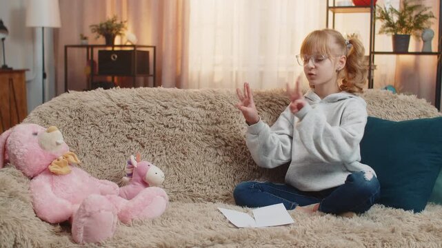 Teenager girl places toys plush figures in front at home sofa, seriously reciting repeats simple math arithmetic. Child kid occasionally giggles laughs amused by own mock authority, silly teacher role