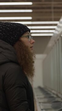Woman Searches Groceries. Female Shopper Wearing Gloves Carefully Examines Product Labels In Cold Storage. Woman In Outerwear Scrutinizes Package Labels And Opens Freezer Doors With Focus