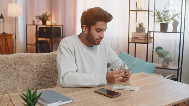 Young Indian man holds cash in dollars, thoughtfully plans expenses using calculator on smartphone, organizing money into separate piles on table in living room at home, managing finances carefully.