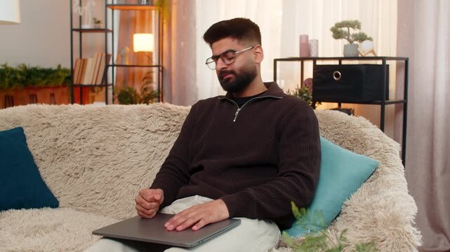 Indian man at home closes laptop angrily, waves hand after internet issue interrupts work. Arabian guy on sofa reacts with visible frustration and disappointment caused by digital connection failure