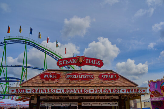 Stuttgart, Germany - May - 06, 2023: Vibrant Stuttgart spring festival scene with STEAK HAUS stall, roller coaster, flags, and sky.