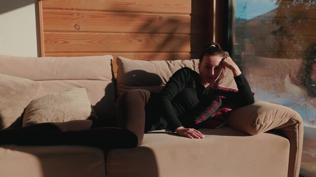 Young woman relaxing on beige sofa near window in wooden cabin interior, cozy lifestyle portrait with warm sunlight, casual home rest, calm female lying on couch in modern rustic living room