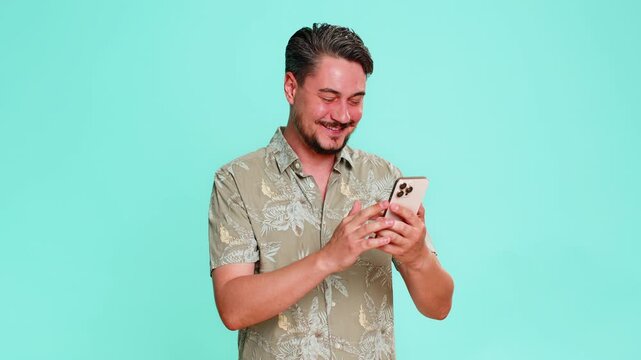 Middle-aged man joyfully laughing, looking at smartphone screen, reacting to funny memes or viral online videos. Young adult guy isolated on blue background enjoying digital humor and entertainment