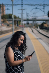 Obraz premium Smiling woman using smartphone while waiting on railway platform, connecting and traveling by public transport