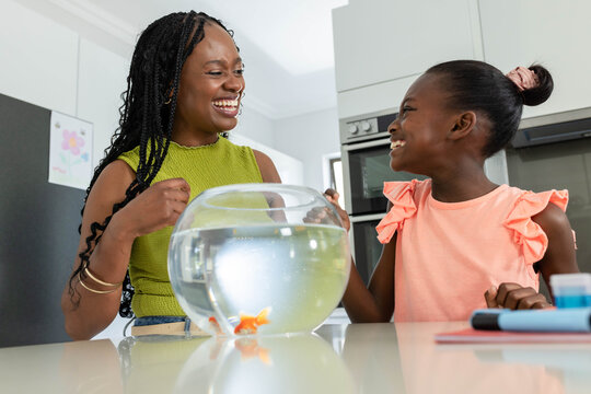 African American mother and daughter in green and pink laughing at kitchen counter with fishbowl