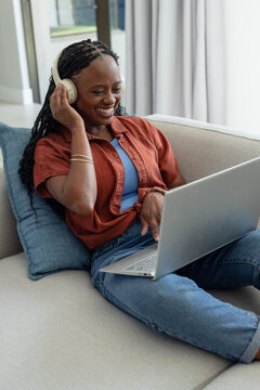 African American woman sitting on sofa at home with blue pillow wearing headphones and using laptop