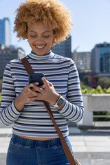 African American woman standing on rooftop wearing striped crop top and jeans, holding smartphone