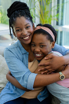 African American mother and teen daughter hugging on sofa by windows, wearing denim and headband