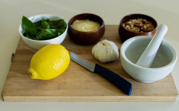 Whole lemon is sitting on cutting board on countertop, with knife, basil, grated cheese and mortar
