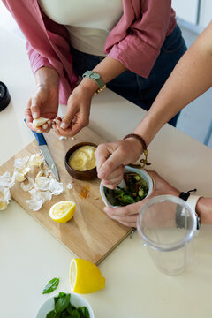 Couple peeling garlic and crushing basil with mortar on wooden cutting board near blender