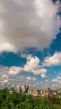 Fluffy white clouds gracefully gliding across a vibrant blue sky over the city skyline of Kyiv, Ukraine, surrounded by lush green trees, creating a serene and picturesque urban landscape