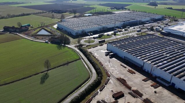 Drone establishing view of a large logistics and industrial warehouse in San Nazzaro, Province of Piacenza, with a full rooftop solar panel system, beside SS10 and surrounded by Po Valley farmland.