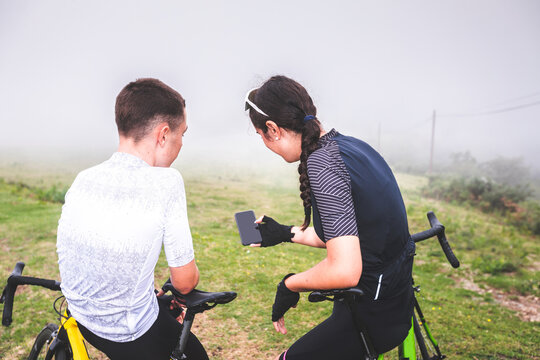 Young woman and man cyclist looking a cellphone wearing cycling cloathes next to a road.