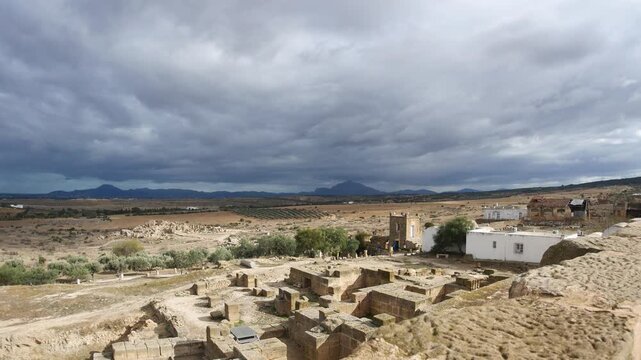 Panorama: Uthina archaeological site Tunisia featuring ancient Roman columns on elevated terrace, wide angle view overlooking countryside beneath dramatic cloudy sky, highlighting classical heritage