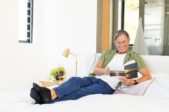 Mature adult male reclining on bed reading paperback with sunset cover in bright bedroom
