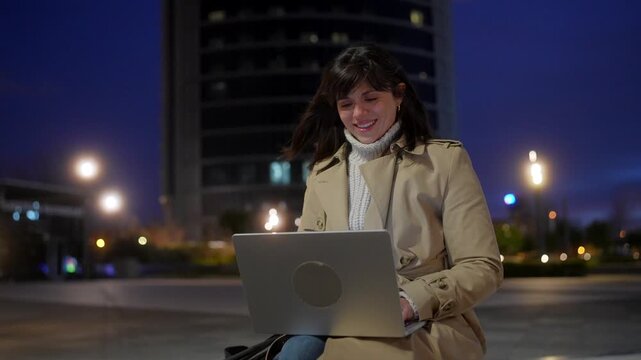 Confident businesswoman smiling while working remotely on her laptop in a modern city square at night, with illuminated buildings and streetlights creating a beautiful bokeh background