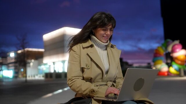 Young businesswoman sitting outdoors in a modern city square at night, smiling and typing on her laptop computer while working remotely, with illuminated buildings in the background