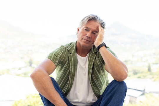 Senior man sitting on ledge, holding head, wearing striped shirt, smartwatch, overlooking suburb
