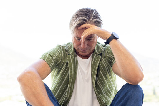 Mature man sitting, bowing head, touching forehead on balcony in green shirt and left-wrist watch