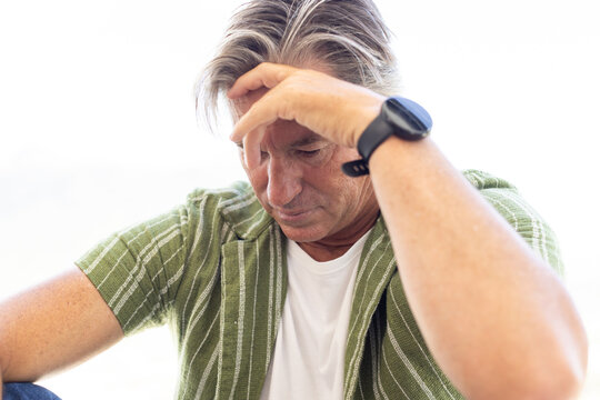 Senior man sitting indoors, holding head with forearm, wearing green shirt over white tee and watch