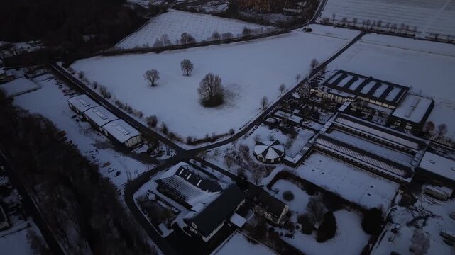 Aerial dusk view of Warsteiner World covered in snow, showing brewery complex surrounded by winter fields and countryside. Industrial architecture meets calm rural landscape in Germany. Tourist spot.