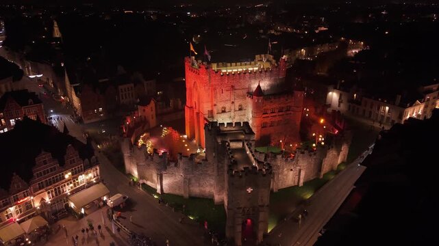 A nighttime drone approach to the Gravensteen (Castle of the Counts) in Ghent, illuminated in a striking red glow during the "Winter Wonder Castle" event, reflecting in the Lieve canal.