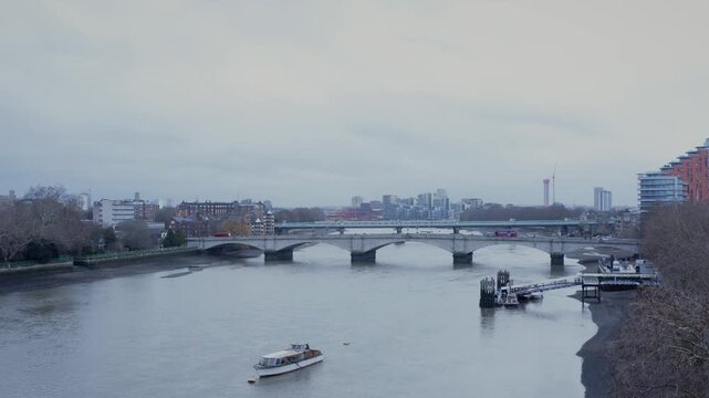 Aerial drone shot panning smoothly across the River Thames, revealing Putney Bridge spanning the river. Natural daylight with gentle water movement below, urban riverside architecture in the backgroun