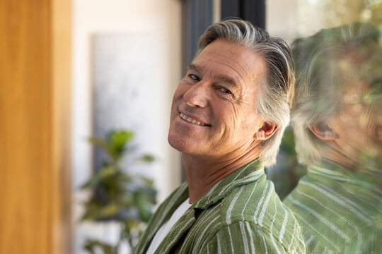 Mature man leaning on glass at home, smiling, in green striped tee, reflection, plant, wall art