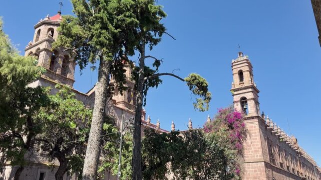 Wide shot of historic Clavijero Cultural Center in Morelia, Michoacan, featuring colonial stone towers and lush green trees under clear blue sky