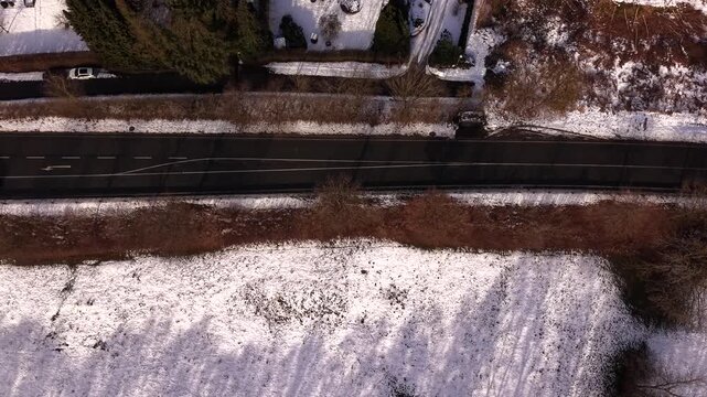 Top down aerial view of cars driving on snow bordered road in winter on sunny day. Traffic movement and road curves in an American landscape from above. Tracking shot. Snowy fields.
